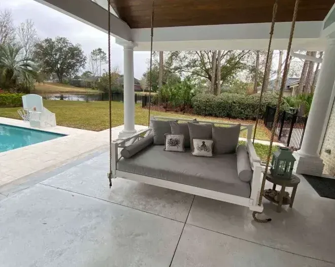 A white porch swing with gray cushions hangs on a patio overlooking a pool and yard. A white porch swing with gray cushions hangs on a patio overlooking a pool and yard.